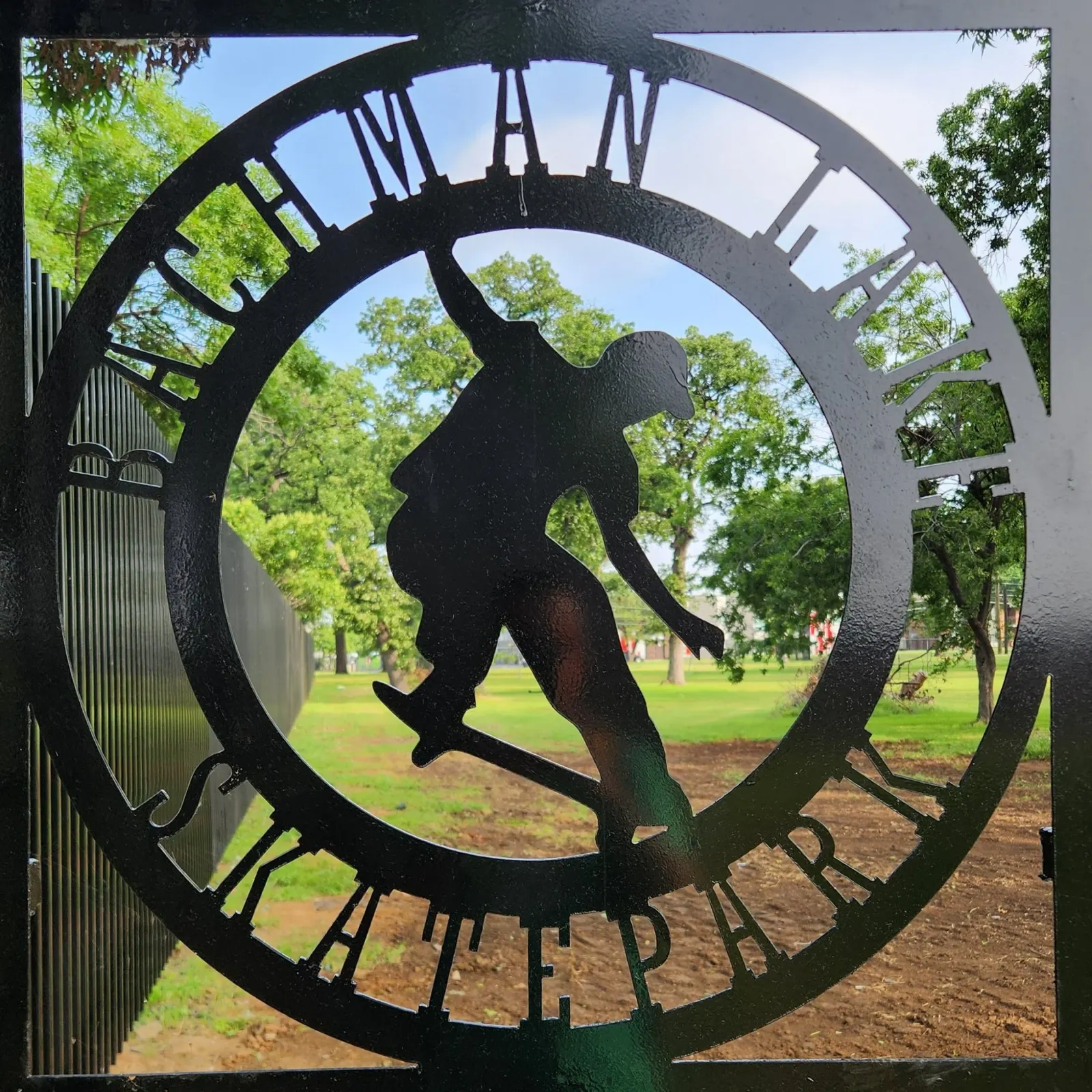 The Bachman Lake Skatepark entrance sculpture: a black steel circle with a skateboarder silhouette cut out of it, park lawn beyond